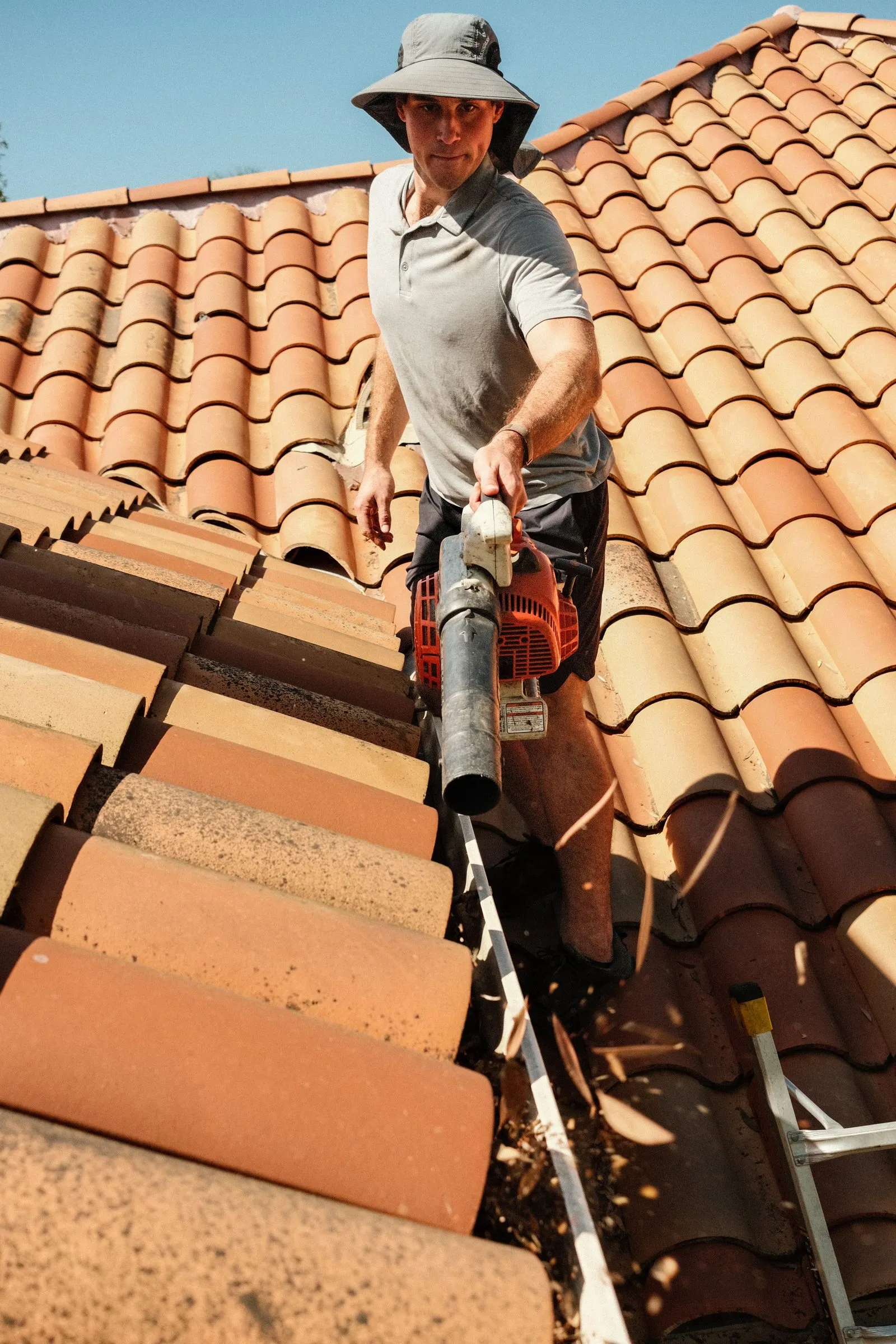 Elliot on a tile roof clearing a gutter with a blower, debris flying